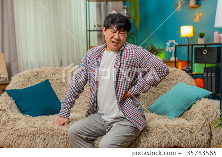 Asian man grimacing from strong back pain, carefully sitting down, painful strain discomfort tension Asian man grimacing from strong back pain, carefully sitting down, painful strain discomfort tension 135783565
