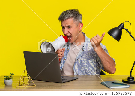 Adult man shouting through megaphone at laptop screen gesturing angrily sitting at working table Adult man shouting through megaphone at laptop screen gesturing angrily sitting at working table 135783578