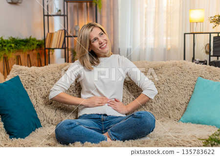 Adult woman holding stomach at home, expressing abdominal pain and discomfort while sitting on sofa Adult woman holding stomach at home, expressing abdominal pain and discomfort while sitting on sofa 135783622