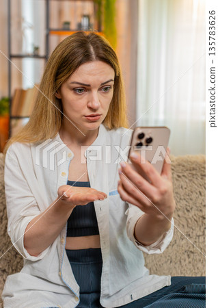 Woman upset reading comments on smartphone at home, reacting emotionally with visible frustration Woman upset reading comments on smartphone at home, reacting emotionally with visible frustration 135783626