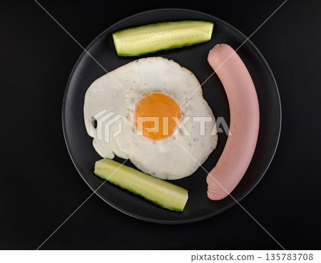A fried egg, sausage and two pieces of cucumber lie on a black plate on a black background A fried egg, sausage and two pieces of cucumber lie on a black plate on a black background 135783708