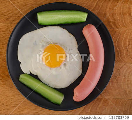 Scrambled eggs, sausage, cucumbers in a black plate on a wooden board Scrambled eggs, sausage, cucumbers in a black plate on a wooden board 135783709