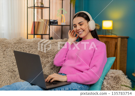 Young woman sitting on sofa wearing headphones listening music while working on laptop with feet up Young woman sitting on sofa wearing headphones listening music while working on laptop with feet up 135783754