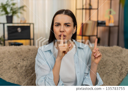 Young woman sitting on sofa making silence gesture placing finger to lips quietly saying shh secret Young woman sitting on sofa making silence gesture placing finger to lips quietly saying shh secret 135783755