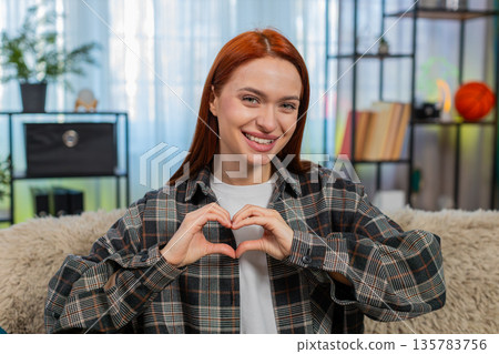Woman sits on sofa making heart shape with fingers smiling showing love and sweet emotional gesture Woman sits on sofa making heart shape with fingers smiling showing love and sweet emotional gesture 135783756