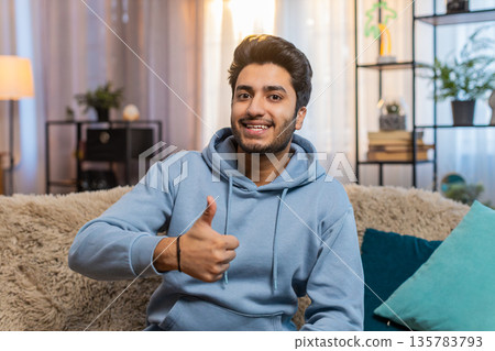 Happy excited Indian man showing thumbs up to camera giving positive feedback sitting on couch Happy excited Indian man showing thumbs up to camera giving positive feedback sitting on couch 135783793