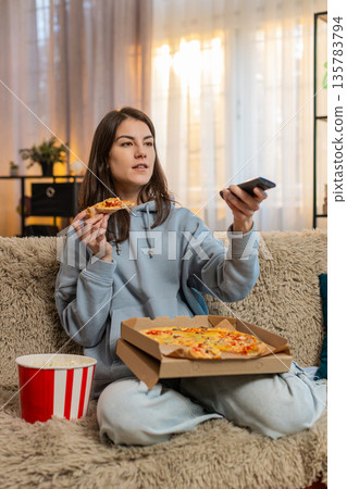 Young woman eating delicious junk food pizza and popcorn sitting on sofa couch watching TV at home 135783794