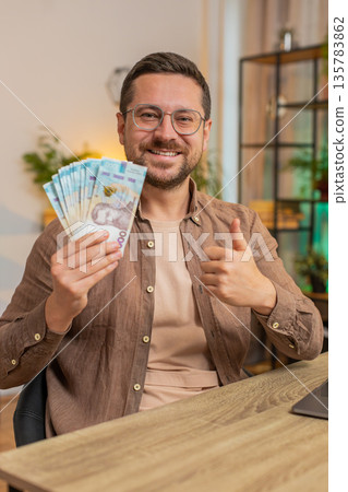 Businessman freelancer holding fan of hryvnias cash showing thumbs up sitting at home office table 135783862
