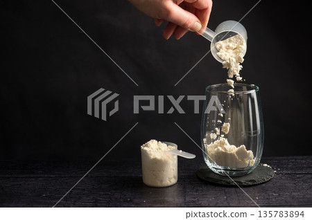 Protein drink. Hand pouring protein powder in a drinking glass. Dark background. Body building, muscle growth and food supplement concept 135783894