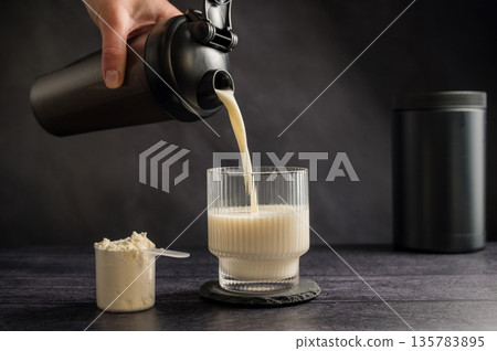 Protein drink. Pouring protein shake in a glass. Dark background. Body building, muscle growth and food supplement concept Protein drink. Pouring protein shake in a glass. Dark background. Body building, muscle growth and food supplement concept 135783895