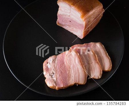 Food, a piece of bacon and cut into slices, on a black plate on a black background Food, a piece of bacon and cut into slices, on a black plate on a black background 135784283