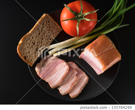 Food, Sliced bacon, tomato green onion and a piece of black bread, on a black plate on a black background Food, Sliced bacon, tomato green onion and a piece of black bread, on a black plate on a black background 135784299