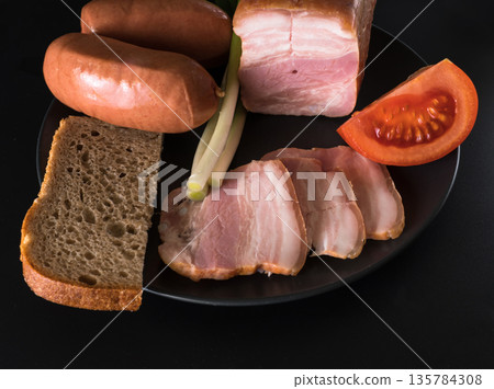 Food, lard, green onions, tomato, black bread, sausages, on a black plate on a black background Food, lard, green onions, tomato, black bread, sausages, on a black plate on a black background 135784308