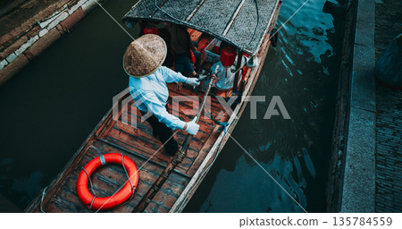 Charming boatman steering through serene waters in a traditional canoe. Zhujiajiao, Shanghai, China Charming boatman steering through serene waters in a traditional canoe. Zhujiajiao, Shanghai, China 135784559