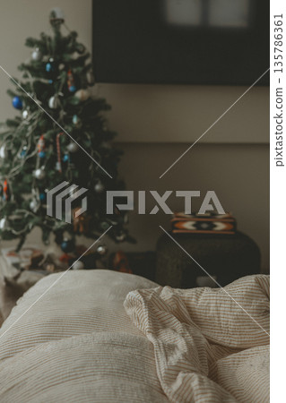 Cozy home bedroom with an unmade bed and soft bedding, creating relaxed homy atmosphere. Decorated Christmas tree. Winter holiday spirit. 135786361