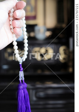 A woman's left hand holding a pearl rosary in front of a Buddhist altar 135787221
