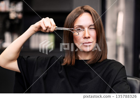 Beautiful female hairstylist looking through hairdressing scissors while sitting in modern hair salon. Professional woman in dark clothes, indoor beauty studio, creative portrait, fashion and style 135788326