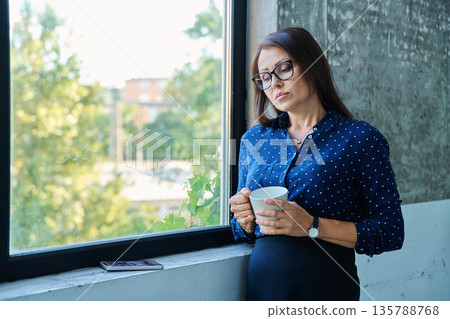 Mature tired, stressed businesswoman standing near window Mature tired, stressed businesswoman standing near window 135788768