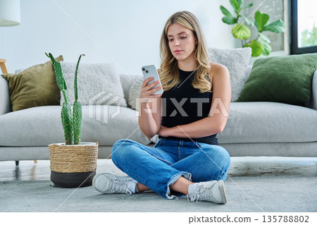 Serious young woman with smartphone sitting at home near sofa Serious young woman with smartphone sitting at home near sofa 135788802