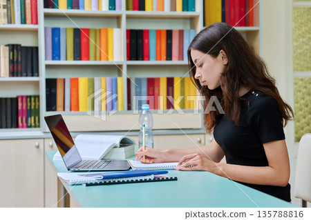 Teenage high school student girl studies in the library with laptop and books 135788816