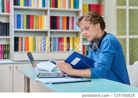 Teenage high school student guy studies in the library with laptop and books Teenage high school student guy studies in the library with laptop and books 135788828