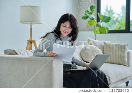 Young smiling student girl sitting at home on couch with laptop, reading papers Young smiling student girl sitting at home on couch with laptop, reading papers 135788920