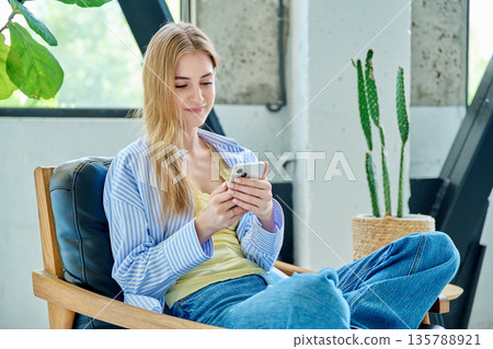 Young smiling woman sitting at home on chair using smartphone 135788921