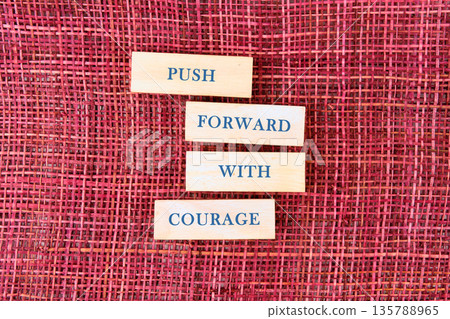 Four wooden blocks are stacked atop one another. Each block features a word, spelling out the phrase PUSH FORWARD WITH COURAGE. They rest on a deep ruby-colored woven surface Four wooden blocks are stacked atop one another. Each block features a word, spelling out the phrase PUSH FORWARD WITH COURAGE. They rest on a deep ruby-colored woven surface 135788965