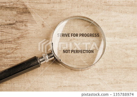 A magnifying glass focuses on the words strive for progress not perfection printed on a textured wooden background. Its a reminder to value improvement over faultless results 135788974