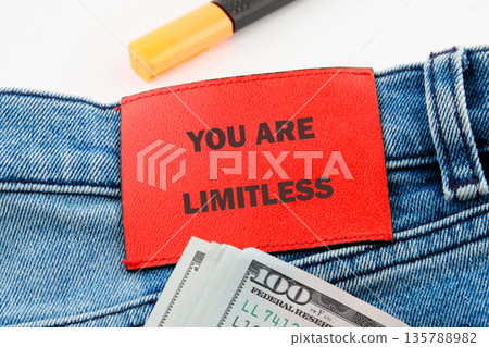 A close-up showcases a stack of one hundred United States Dollar bills near blue jeans with a bright red patch saying YOU ARE LIMITLESS. A yellow highlighter rests nearby A close-up showcases a stack of one hundred United States Dollar bills near blue jeans with a bright red patch saying YOU ARE LIMITLESS. A yellow highlighter rests nearby 135788982