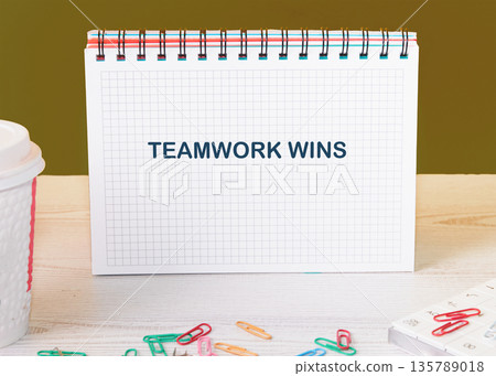 A white spiral notebook displays the capitalized phrase TEAMWORK WINS on grid paper A white spiral notebook displays the capitalized phrase TEAMWORK WINS on grid paper 135789018