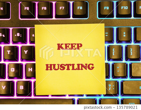 Yellow sticky note with KEEP HUSTLING in red bold text rests on a black computer keyboard. The keys glow with bright purple and blue backlighting, creating an inspiring work scene 135789021