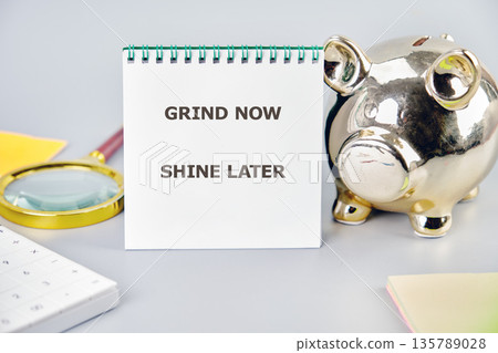 A white spiral notebook displays the text GRIND NOW SHINE LATER positioned beside a gleaming golden piggy bank 135789028