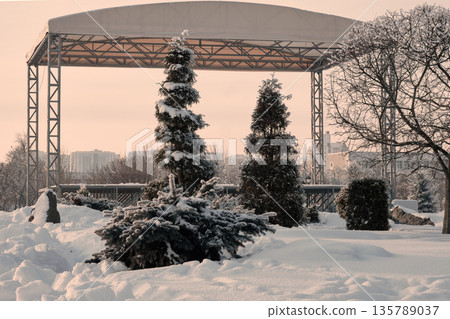 Serene winter landscape in a park with fresh snow covering ground and evergreen trees. A large outdoor metal stage structure dominates the background Serene winter landscape in a park with fresh snow covering ground and evergreen trees. A large outdoor metal stage structure dominates the background 135789037
