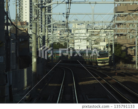 一列JR通勤列車沿著鐵軌行駛，背景是林立的城市建築。這是一幅典型的城市鐵路景象，架空電線錯綜複雜，鐵軌彎曲蜿蜒。 135791133