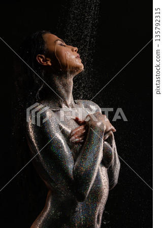 Ethereal woman with glitter body art standing under falling water on a black background Ethereal woman with glitter body art standing under falling water on a black background 135792315
