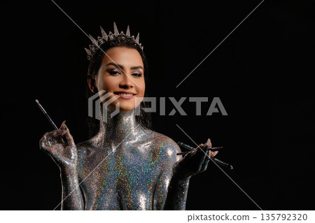 Smiling woman in crown with holographic silver glitter body art holding makeup brushes. 135792320
