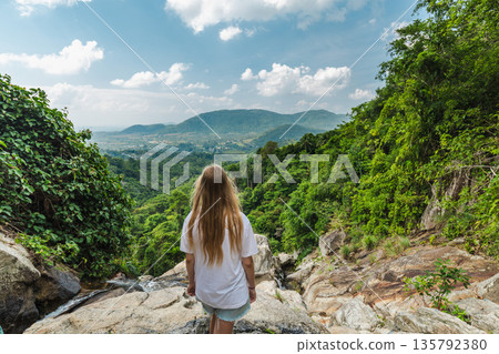 Young woman standing on rocky cliff overlooking lush jungle valley and distant hills near Samui waterfall in Thailand under bright blue sky and tropical landscape atmosphere 135792380