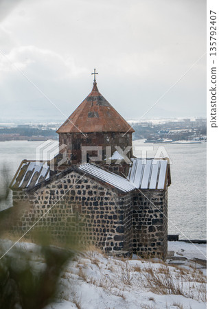 Stone church of Sevanavank Monastery stands above snowy shore of Lake Sevan in Armenia, dark volcanic masonry contrasts with pale winter sky 135792407