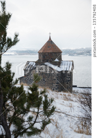 Stone church of Sevanavank Monastery stands above snowy shore of Lake Sevan in Armenia, dark volcanic masonry contrasts with pale winter sky 135792408