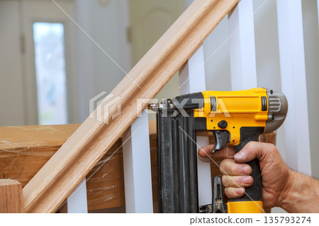 Carpenter uses nail gun to attach wooden railing inside house, tools materials are visible Carpenter uses nail gun to attach wooden railing inside house, tools materials are visible 135793274