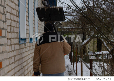 A man carries a snow shovel on his shoulder as he walks alongside a brick house in a snowy yard preparing to remove fresh snow from the area A man walks through the yard with a shovel to remove snow. A man carries a snow shovel on his shoulder as he walks alongside a brick house in a snowy yard preparing to remove fresh snow from the area A man walks through the yard with a shovel to remove snow. 135793765