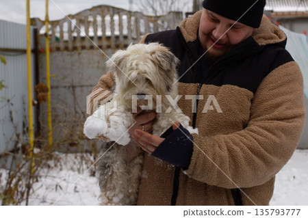 A man smiles, holds his fluffy white dog with snow on his fur and paws, sharing playful, affectionate winter moments outdoors in warm clothes and a hat. A large amount of snow stuck to the dog's paws. A man smiles, holds his fluffy white dog with snow on his fur and paws, sharing playful, affectionate winter moments outdoors in warm clothes and a hat. A large amount of snow stuck to the dog's paws. 135793777
