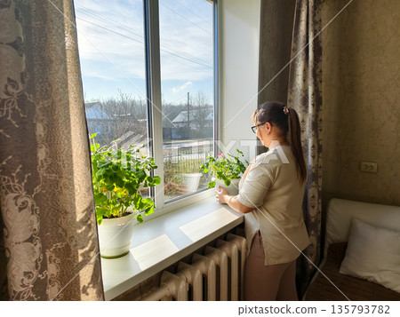 Woman standing by a sunlit window in her cozy apartment, holding a potted plant on the windowsill and gazing out thoughtfully, enjoying a quiet, peaceful moment indoors. 135793782