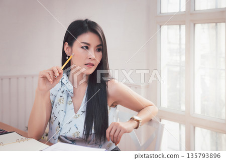 Charming young Asian woman holding a pencil while thinking when looking outside window in office. Charming young Asian woman holding a pencil while thinking when looking outside window in office. 135793896