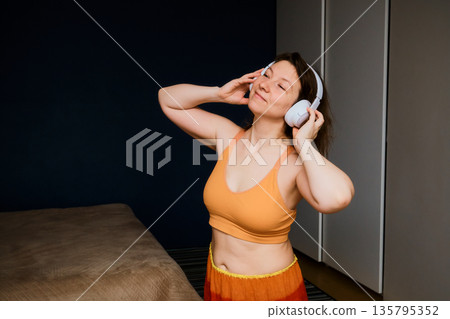 Woman closing eyes listening to music through headphones in bedroom Woman closing eyes listening to music through headphones in bedroom 135795352