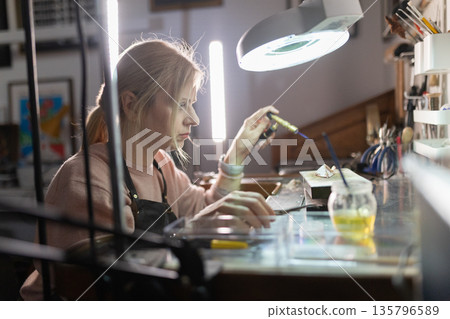 Woman soldering metal artwork in jewelry workshop Woman soldering metal artwork in jewelry workshop 135796589
