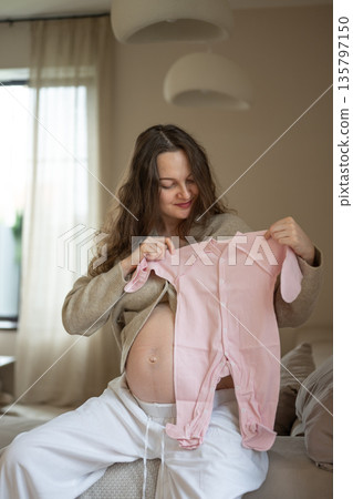 Pregnant woman holding pink baby clothes sitting on sofa 135797150