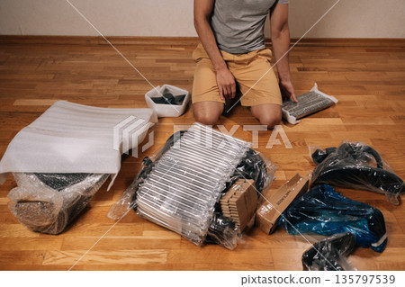 Cropped shot of man kneeling on wooden floor unpacking parts and tools, assembling a new ergonomic office chair at home, following instructions for diy remote-work setup project. Cropped shot of man kneeling on wooden floor unpacking parts and tools, assembling a new ergonomic office chair at home, following instructions for diy remote-work setup project. 135797539