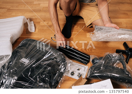 Top view of contractor male unboxing and assembling new flat-pack office chair for home office, carefully unpackaging components on hardwood floor, engaging in diy project. Top view of contractor male unboxing and assembling new flat-pack office chair for home office, carefully unpackaging components on hardwood floor, engaging in diy project. 135797540